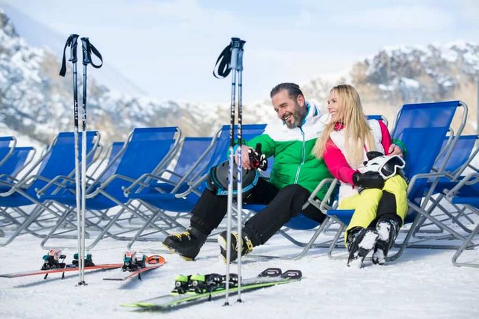 Paar in Skikleidung entspannt sich lachend in blauen Liegestühlen im Schnee mit Blick auf die verschneiten Alpen – Skier und Stöcke im Vordergrund.